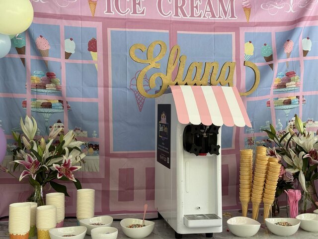 Bash soft serve machine under a pink and white striped awning at a birthday party, with cones, cups and toppings laid out