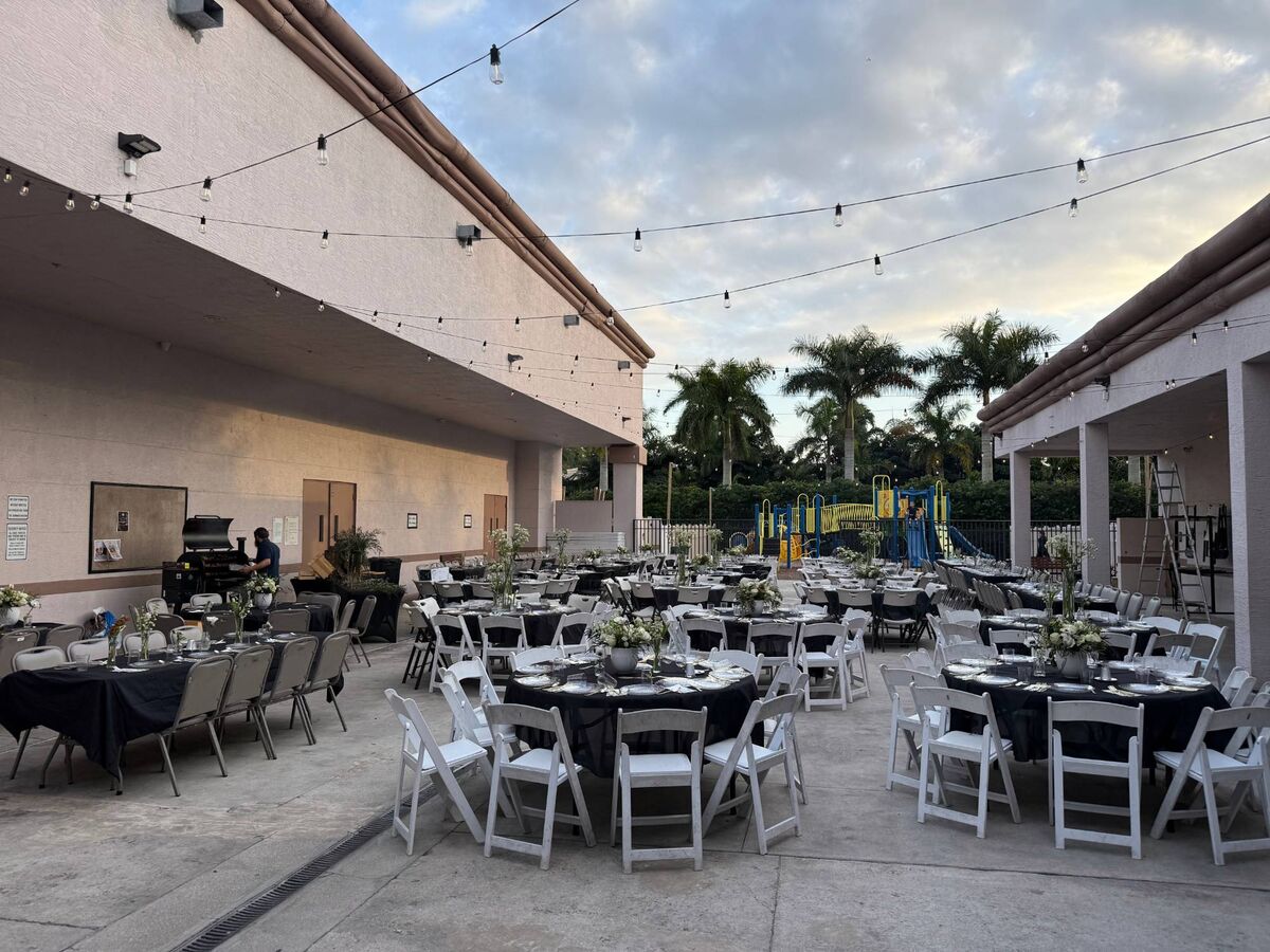 Round tables with black linens and white folding chairs set for an evening dinner in a courtyard